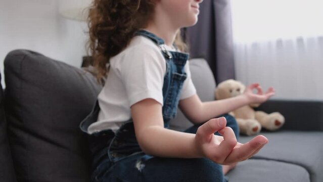 Selective focus of calm little girl child sitting cross-legged on couch in lotus pose with folded fingers, harmony, enjoy mindfulness inner harmony, meditation yoga time. Mental health care concept