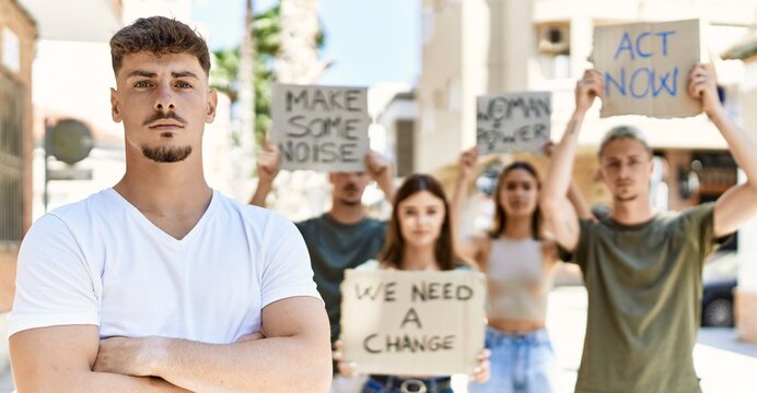 Young hispanic activist man with arms crossed gesture standing with a group of protesters holding banner protesting at the city.