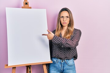 Beautiful hispanic woman pointing to painter easel stand skeptic and nervous, frowning upset...
