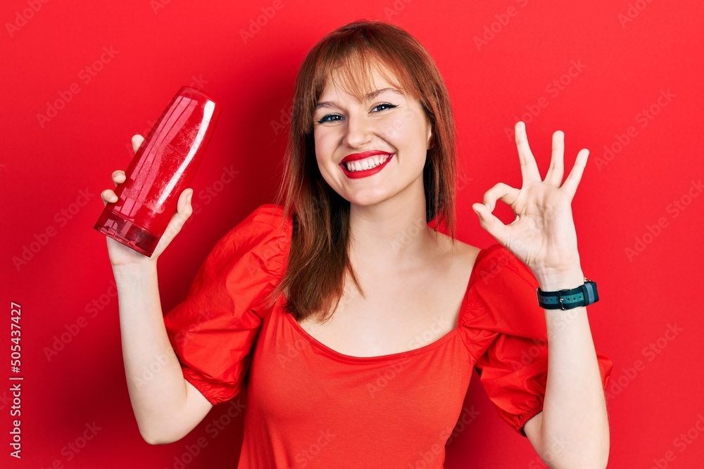 Poster Redhead young woman holding shampoo bottle doing ok sign with fingers, smiling friendly gesturing excellent symbol