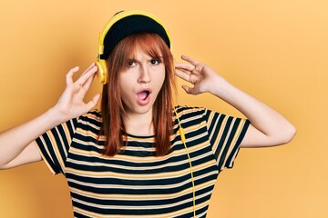 Fototapeta na wymiar Redhead young woman listening to music using headphones in shock face, looking skeptical and sarcastic, surprised with open mouth