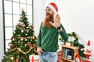 Redhead man with long beard wearing christmas hat by christmas tree waiving saying hello happy and smiling, friendly welcome gesture