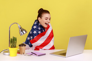 Happy woman employee sitting wrapped in USA flag, shouting for joy in office workplace, celebrating labor day or US Independence day. Indoor studio studio shot isolated on yellow background.
