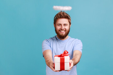 Portrait of handsome bearded angelic man with holy nimbus and pleasant toothy smile giving gift...