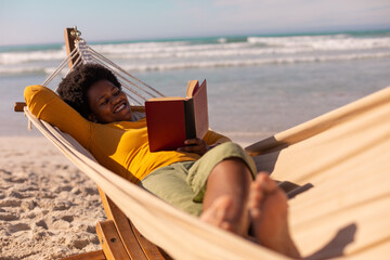 Smiling african american mature woman reading book while lying on hammock against sea in summer