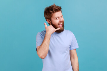 Give me a call. Portrait of positive optimistic bearded man smiling at camera and winking eye, making call me gesture with fingers, flirting. Indoor studio shot isolated on blue background. - obrazy, fototapety, plakaty