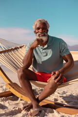 Fototapeta na wymiar Portrait of bearded african american senior man sitting on hammock against blue sky in summer