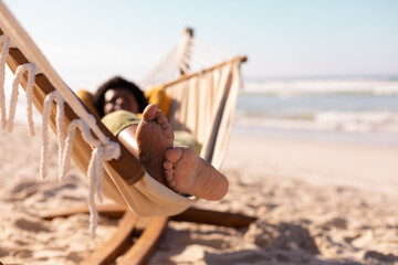 African american mature woman with sand on feet lying on hammock against sea and sky in summer