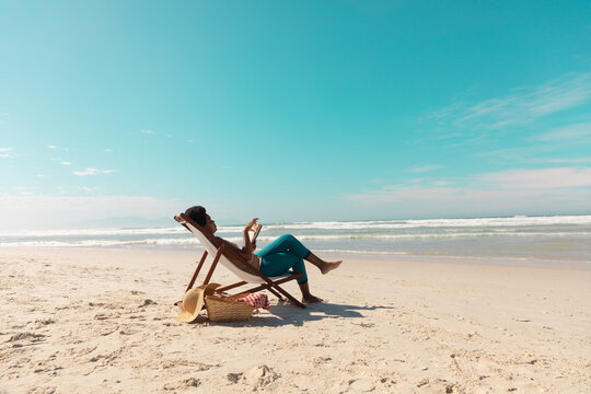 African American Mature Woman Using Digital Tablet Relaxing On Deckchair At Beach Against Sky