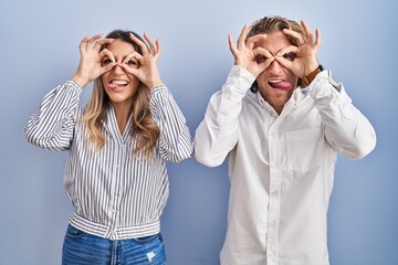 Young couple standing over blue background doing ok gesture like binoculars sticking tongue out, eyes looking through fingers. crazy expression.