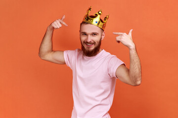 Portrait of smiling handsome bearded man pointing fingers on golden crown on his head, showing his authority, wearing pink T-shirt. Indoor studio shot isolated on orange background. - obrazy, fototapety, plakaty
