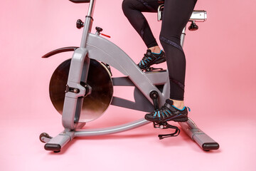 Fototapeta na wymiar Portrait of the bike simulator and women's legs in black sneakers and leggins, cardio workout, training. Indoor studio shot isolated on pink background.
