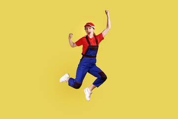 Full length portrait of extremely happy worker woman jumping and clenched fists, celebrating long awaited day off, wearing overalls and red cap. Indoor studio shot isolated on yellow background.