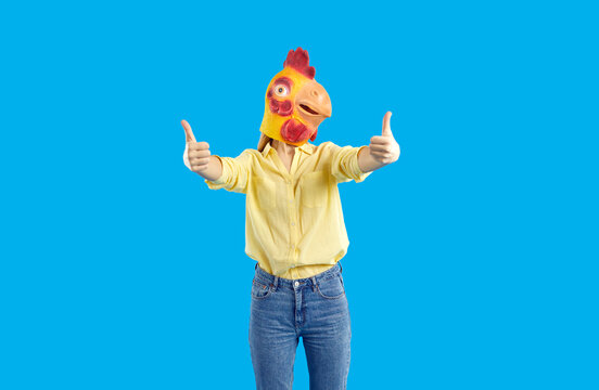 Creative advertising. Cheerful funny and humorous woman in rubber mask of chicken shows thumbs up on blue background. Woman in jeans and yellow shirt wears absurd animal mask and recommends something.