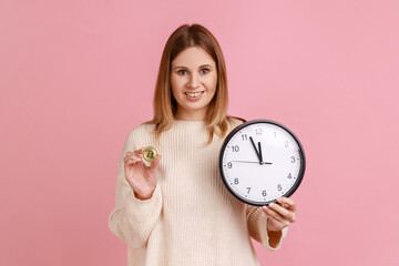 Portrait of positive rich blond woman holding golden coin of crypto currency and wall clock, time...
