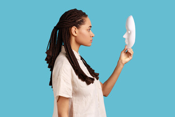 Side view of confident serious woman with dreadlocks holding and looking at white mask with attentive look, trying to understand hiding personality. Indoor studio shot isolated on blue background. - obrazy, fototapety, plakaty
