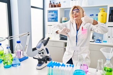 Middle age blonde woman working at scientist laboratory doing thumbs up and down, disagreement and agreement expression. crazy conflict