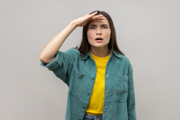 Curious young woman keeping palm over head and looking attentively far away, peering with expectation at long distance, wearing casual style jacket. Indoor studio shot isolated on gray background.