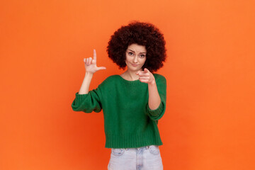 You are loser! Woman with Afro hairstyle wearing green casual style sweater showing loser gesture and pointing finger on you, abuser. Indoor studio shot isolated on orange background.