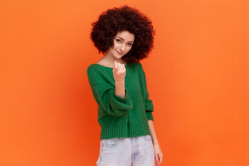 Come here! Woman with Afro hairstyle wearing green casual style sweater making beckoning gesture, inviting to come, flirting and looking playful. Indoor studio shot isolated on orange background.