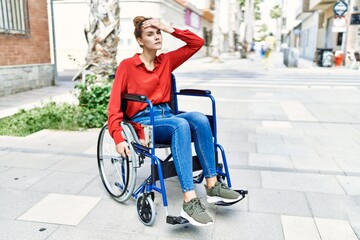 Young brunette woman sitting on wheelchair outdoors stressed and frustrated with hand on head, surprised and angry face