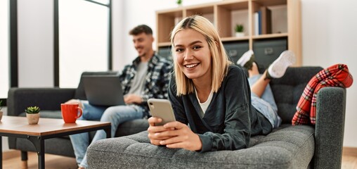 Young caucasian couple smiling happy using laptop and smartphone sitting on the sofa at home.