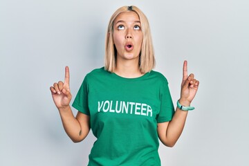 Beautiful blonde woman wearing volunteer t shirt amazed and surprised looking up and pointing with...