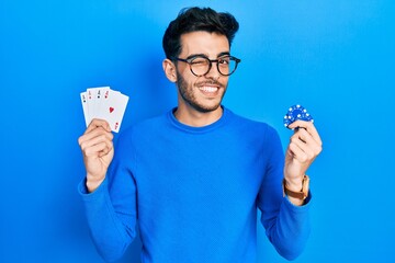 Young hispanic man playing poker holding casino chips and cards winking looking at the camera with...