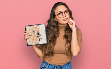 Young hispanic girl holding drauhgts serious face thinking about question with hand on chin, thoughtful about confusing idea