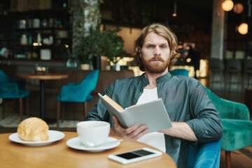 Fototapeta na wymiar Confident young man in casual shirt sitting in armchair and looking away while talking by phone in modern cafe