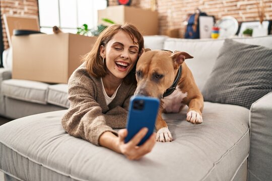 Young caucasian woman make selfie by the smartphone lying on sofa with dog at new home