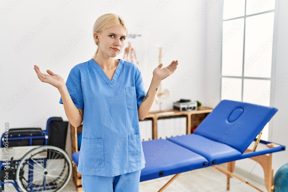 Poster Beautiful caucasian physiotherapist woman working at pain recovery clinic clueless and confused expression with arms and hands raised. doubt concept.