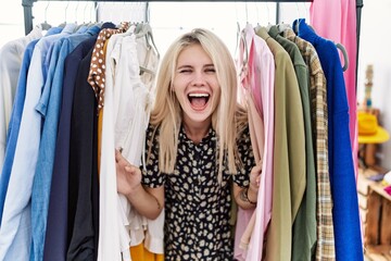 Naklejka na ściany i meble Young blonde woman searching clothes on clothing rack celebrating crazy and amazed for success with open eyes screaming excited.