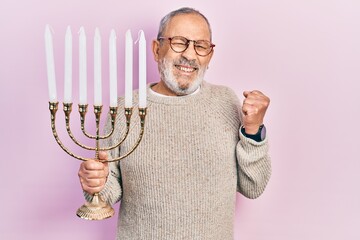 Handsome senior man with beard holding menorah hanukkah jewish candle screaming proud, celebrating...