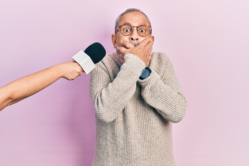 Handsome senior man with beard being interviewed by reporter holding microphone shocked covering...