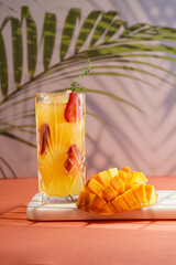 tall cocktail glass filled with yellow juice and fresh strawberries and cut in cubes mango half on tropical palm tree background