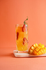 tall cocktail glass filled with yellow juice and fresh strawberries and cut in cubes mango half on tropical orange background