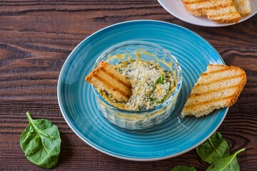 Hot appetizer, spinach and cream cheese dip in a transparent form on a blue plate on a brown wooden background. Served with toasted wheat bread.