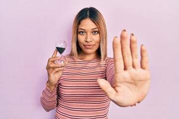 Beautiful hispanic woman holding sand clock with open hand doing stop sign with serious and confident expression, defense gesture