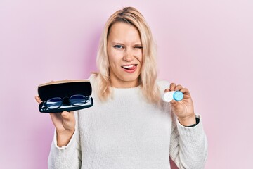 Beautiful caucasian blonde woman holding glasses and contact lenses winking looking at the camera...