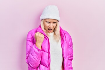 Beautiful caucasian blonde woman wearing wool hat and winter coat angry and mad raising fist frustrated and furious while shouting with anger. rage and aggressive concept.