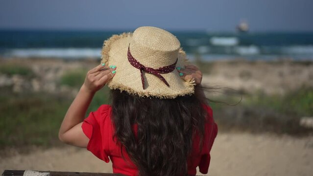 Back view unrecognizable young woman putting on straw hat sitting on bench admiring Mediterranean sea. Caucasian female millennial tourist enjoying summer day on Cyprus outdoors. Slow motion