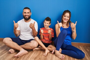 Family of three sitting on the floor at home success sign doing positive gesture with hand, thumbs...