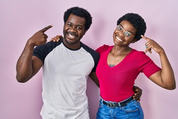 Young african american couple standing over pink background smiling pointing to head with both...