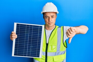 Handsome young man holding photovoltaic solar panel with angry face, negative sign showing dislike...