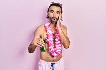 Young hispanic man wearing swimsuit and hawaiian lei drinking tropical cocktail afraid and shocked,...