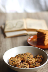 Bowl of chocolate chip cookies, plate of biscuits, open books and reading glasses on the table. Selective focus.