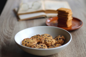 Bowl of chocolate chip cookies, plate of biscuits, open books and reading glasses on the table. Selective focus.