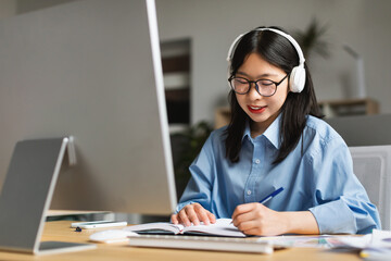 Woman sitting at desk, using computer and writing in notebook