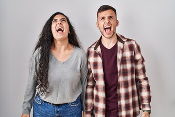Young hispanic couple standing over white background angry and mad screaming frustrated and...